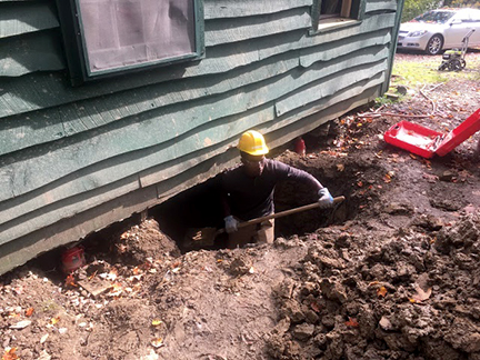 student wearing hard hat digging a hole underneath the house