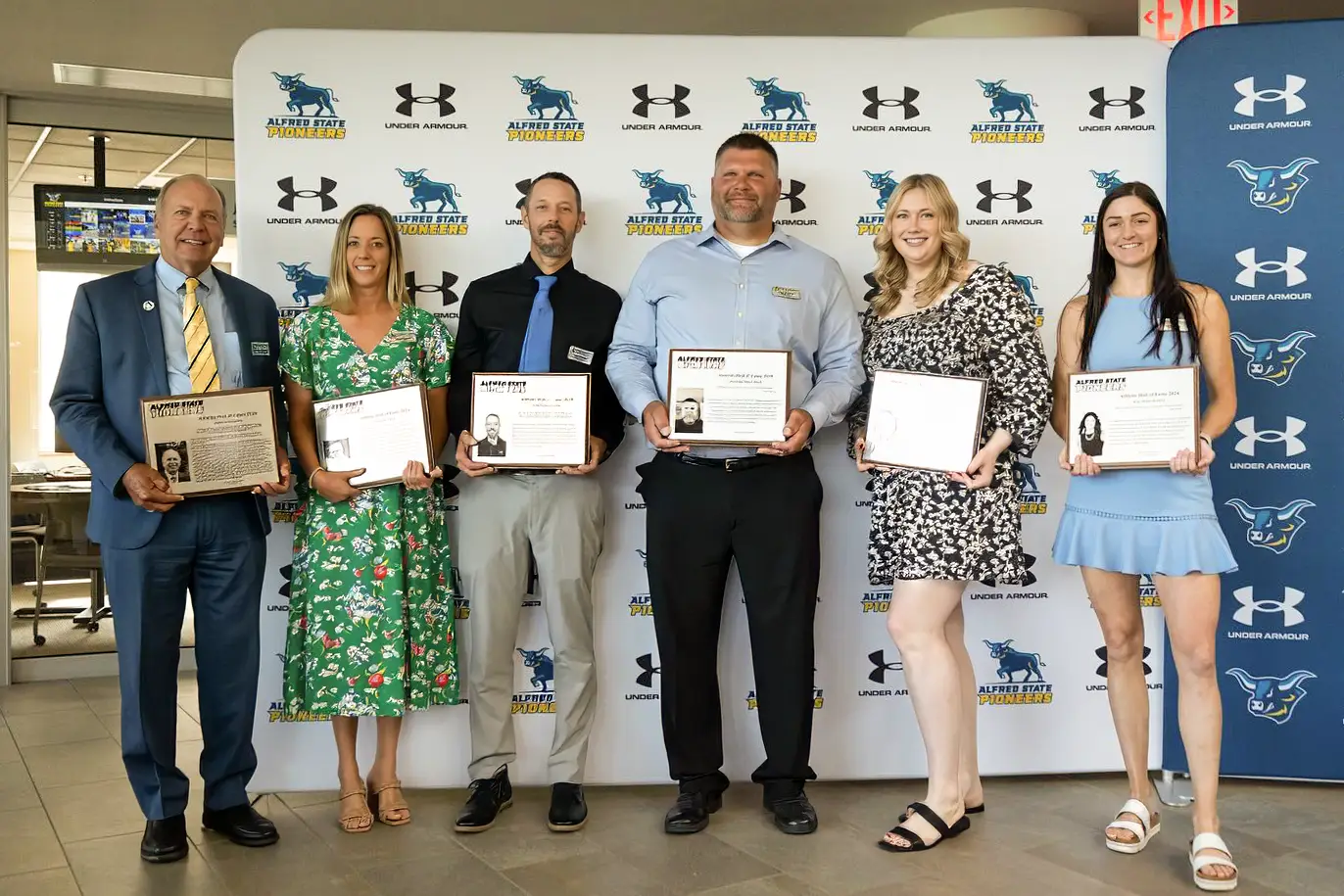 Six individuals pose for a group photo, each holding a plaque in recognition of their achievements. The backdrop features the Alfred State Pioneers logo and Under Armour branding, creating a formal and celebratory atmosphere.