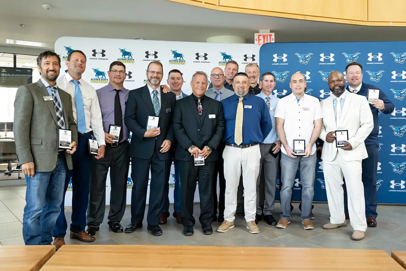 A group of well-dressed individuals poses together indoors, each holding a plaque recognizing their achievements. The backdrop features Alfred State Pioneers and Under Armour logos, emphasizing the formal and celebratory nature of the event.