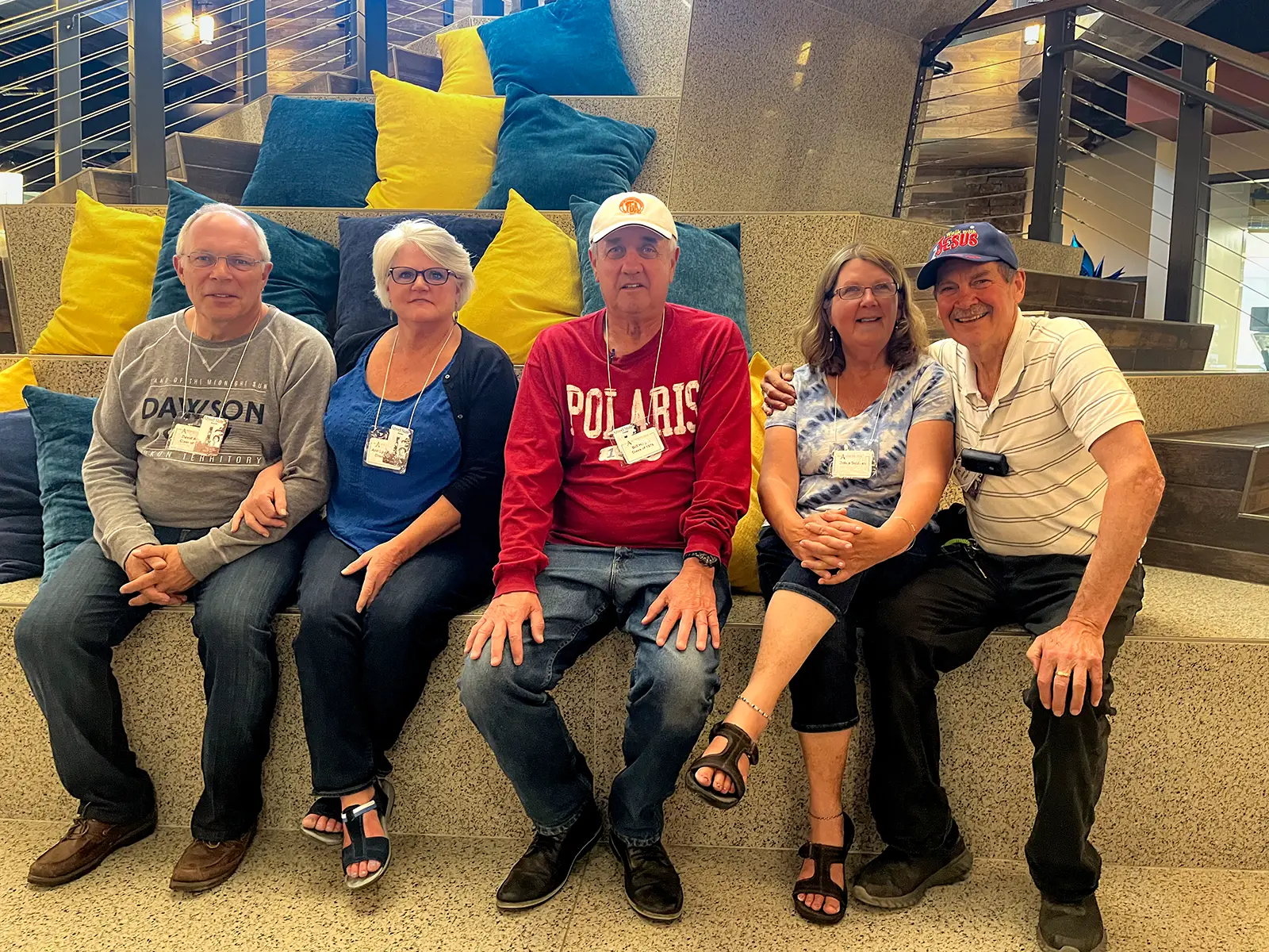 Five individuals sit together on steps decorated with blue and yellow cushions, smiling for a group photo. The setting is indoors, featuring a modern design with railings and bright lighting in the background. Each person wears a name tag, suggesting a reunion or event.
