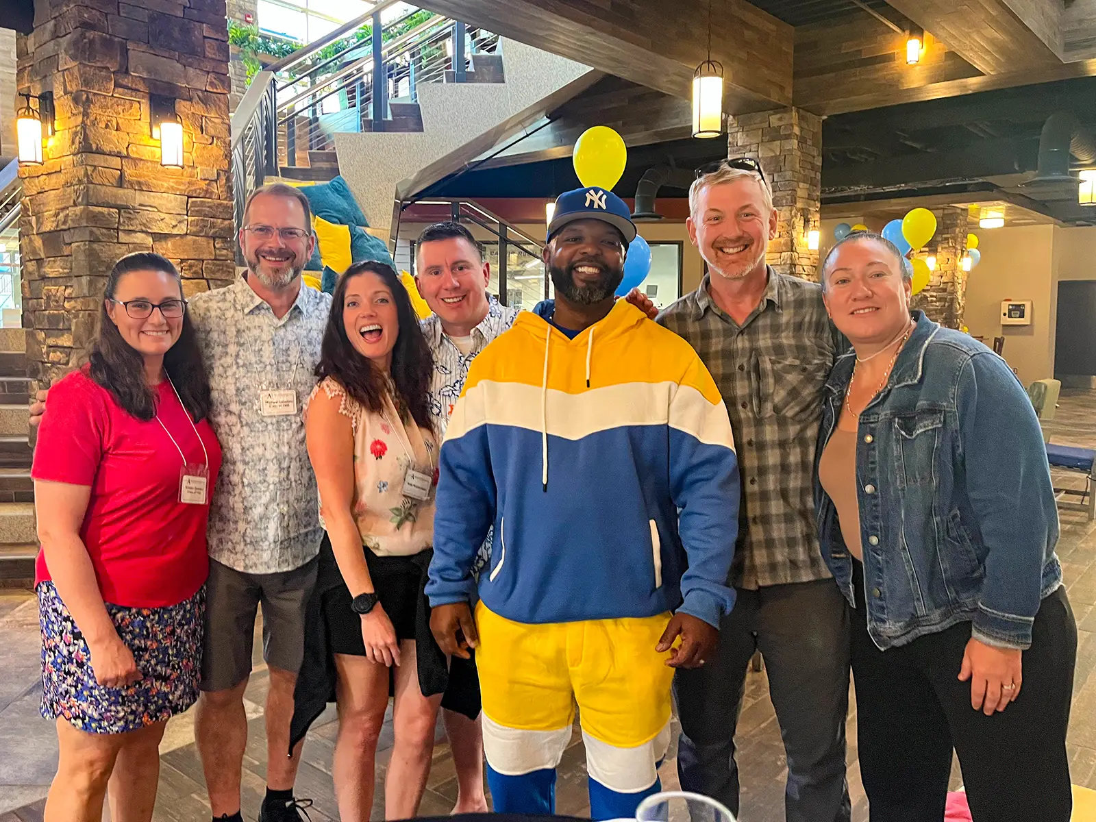 A group of seven individuals poses together indoors, smiling and enjoying a festive event. The setting features bright lighting, stone pillars, and blue and yellow balloons, creating a celebratory atmosphere. Name tags indicate a formal or reunion gathering.