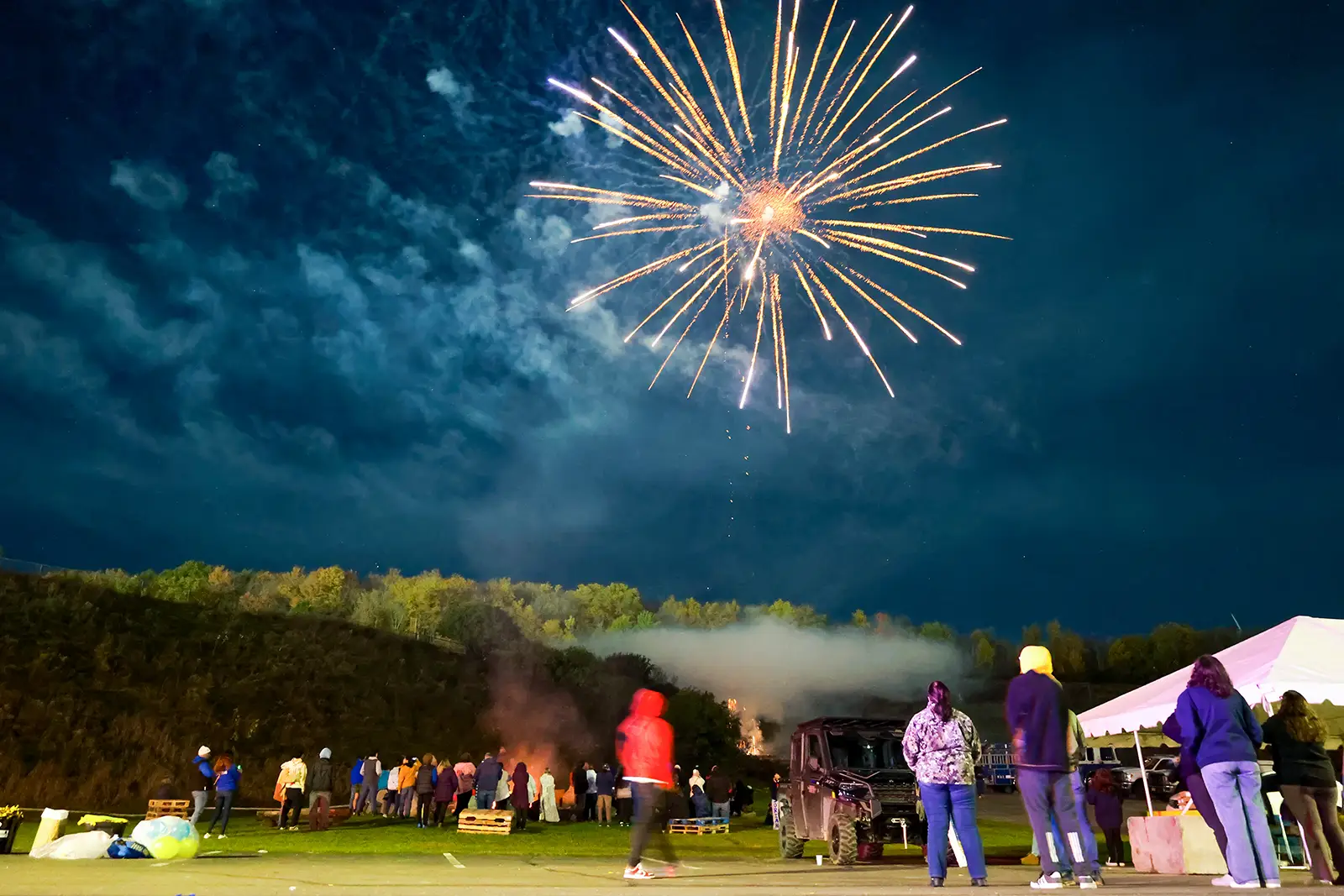 Fireworks light up the night sky above a group of people gathered outdoors. The scene includes a bonfire, a parked vehicle, and a tent, with the backdrop of a wooded hillside. The vibrant display and festive atmosphere highlight the celebration.