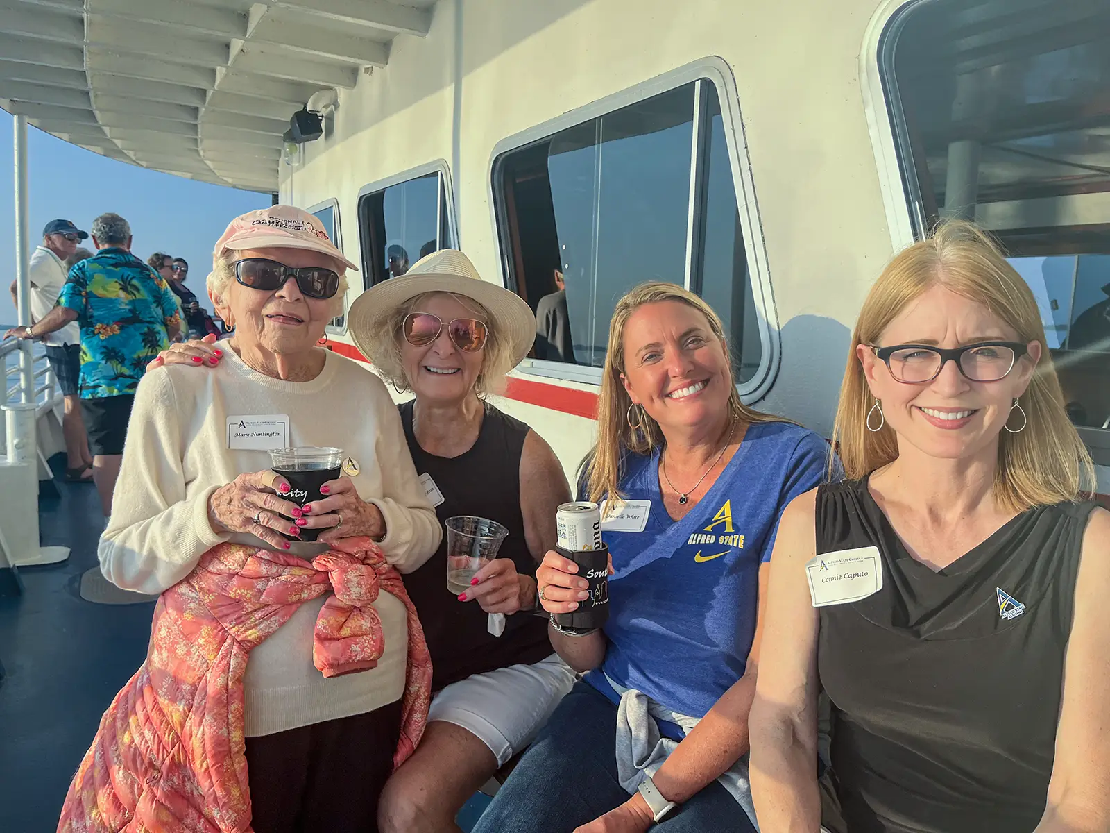 Four women smile for a photo aboard a boat on a sunny day. They are seated near a window, holding drinks, and dressed casually, with one wearing a sunhat and another in a cap. The boat’s deck and other passengers enjoying the outing are visible in the background.