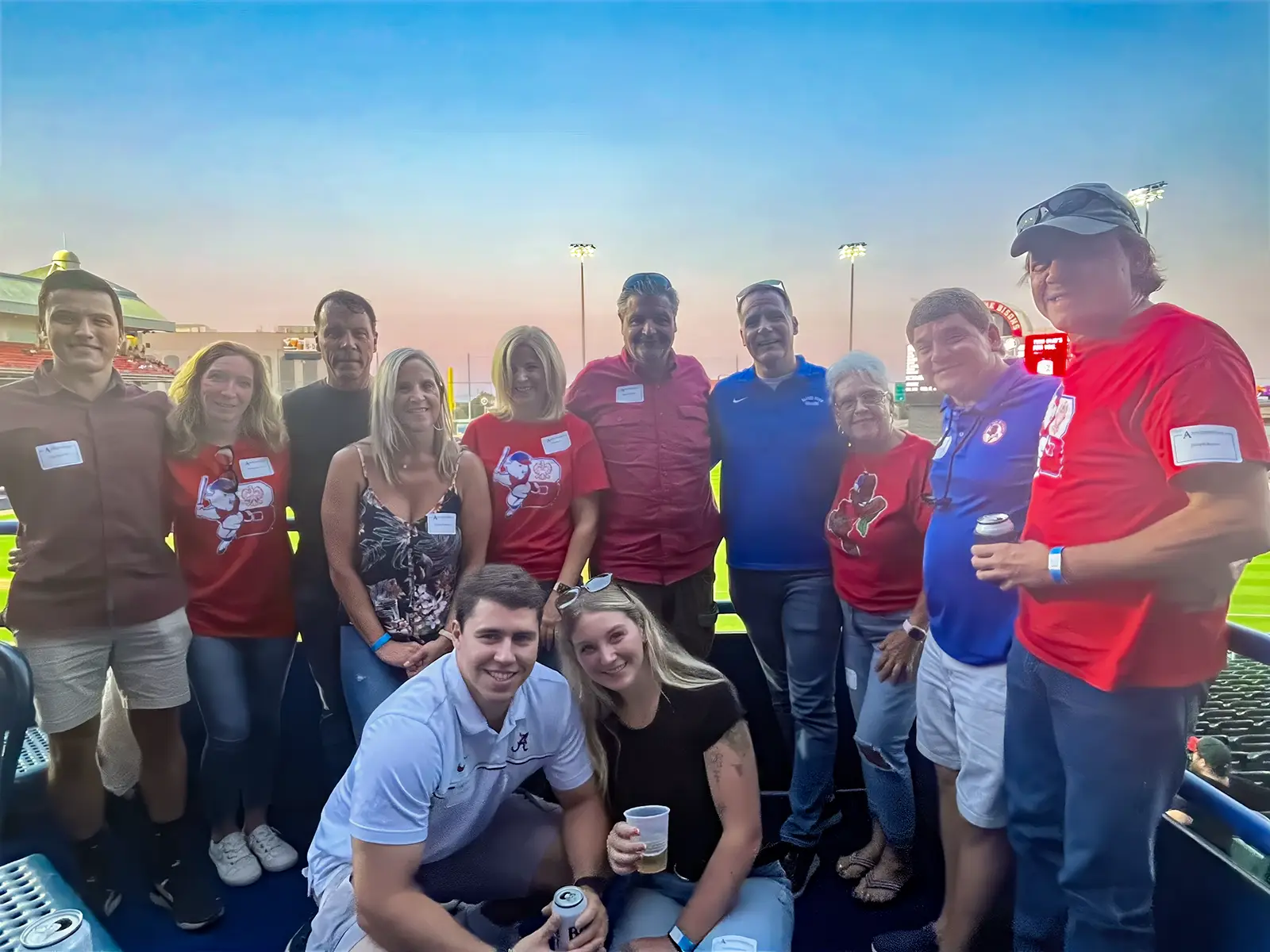 A group of people gathers for a photo at an outdoor event during sunset. Some wear red shirts with festive graphics, and others are dressed casually. The background features a brightly lit sports field with stadium lights and seating.