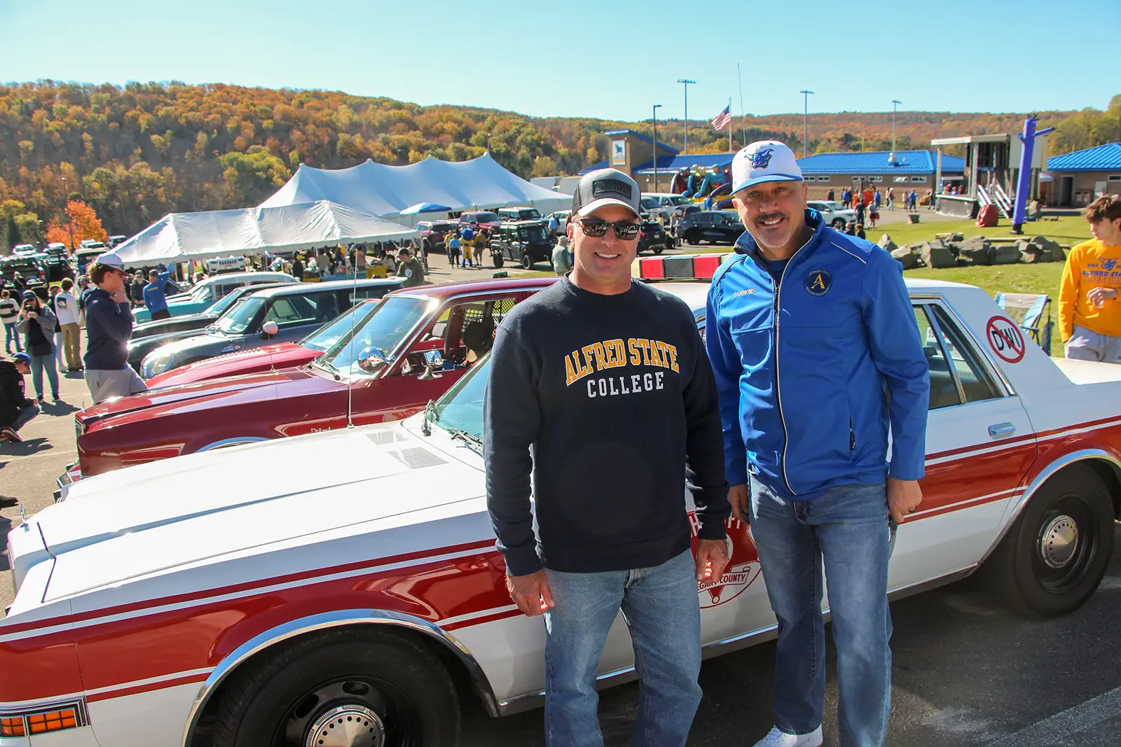 Two men stand smiling in front of a row of vintage cars at an outdoor event. One wears an Alfred State College sweatshirt, and the other wears a blue Alfred State jacket and cap. The background features tents, a stage, and autumn foliage under a clear sky.