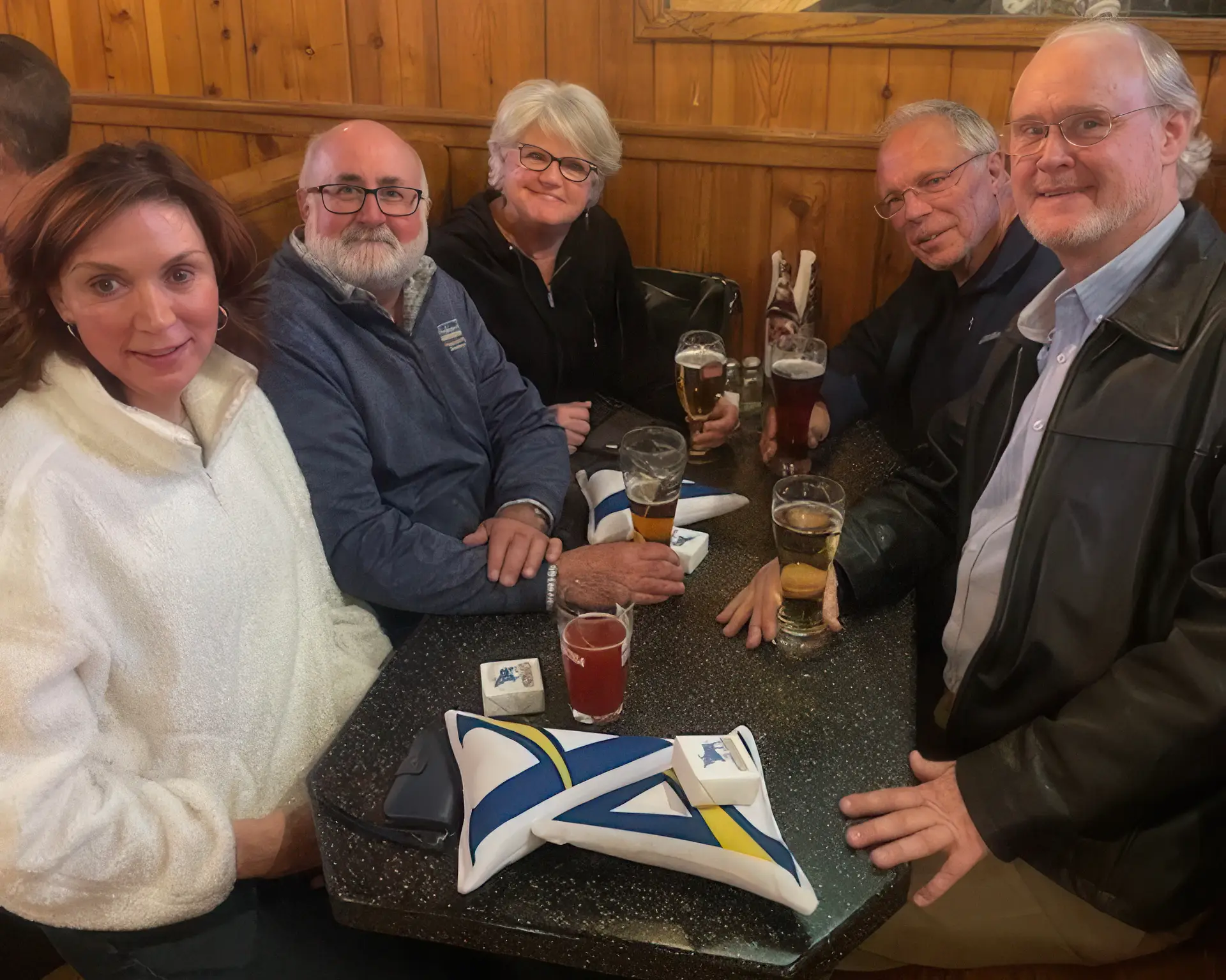 Five individuals sit together at a table in a cozy wooden-paneled setting, enjoying drinks and conversation. The table features small branded items, adding a touch of formality to the relaxed atmosphere.
