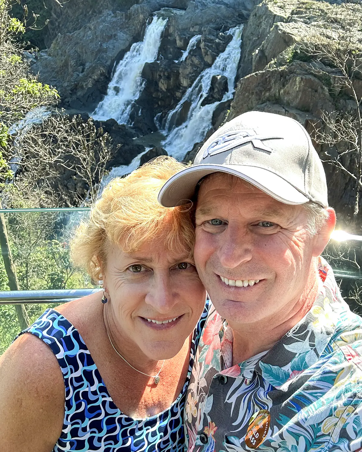 A smiling couple takes a selfie in front of a scenic waterfall. The woman wears a patterned dress, and the man wears a floral shirt and a cap. The lush surroundings and cascading water create a picturesque backdrop.