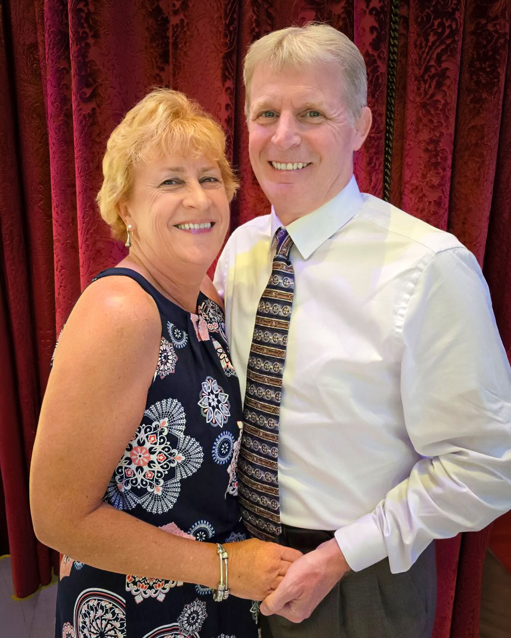 A smiling couple poses together indoors in front of rich red curtains. The woman wears a patterned dress, and the man wears a white shirt with a patterned tie. They stand closely, holding hands, radiating happiness.