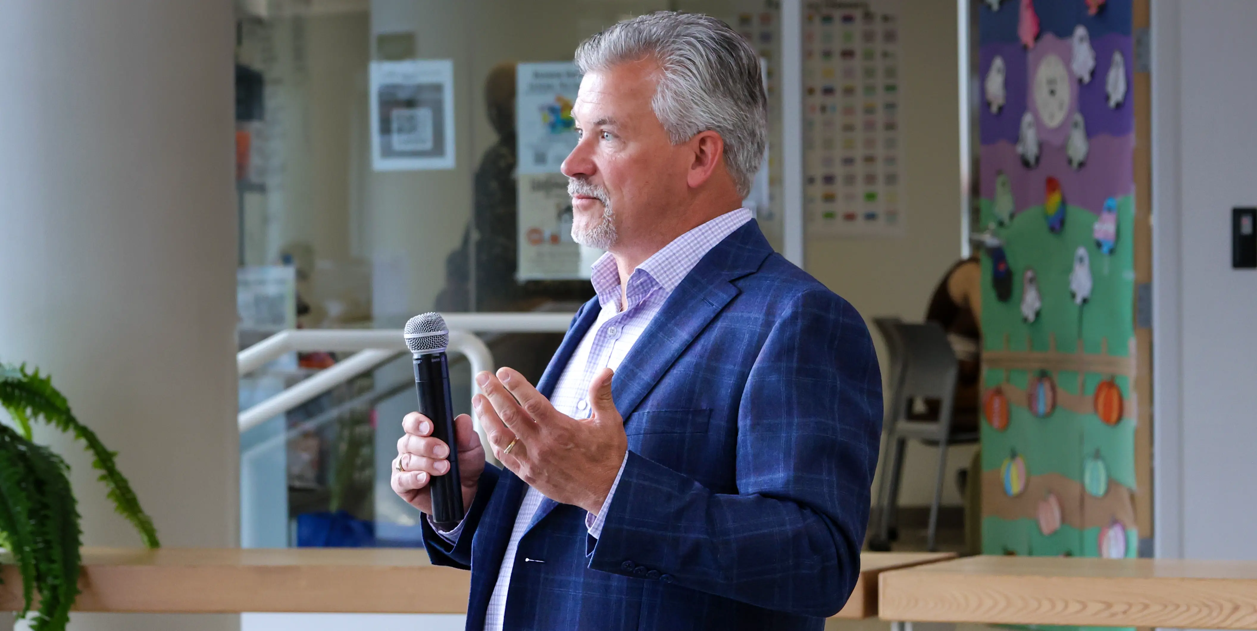 A man in a blue blazer and purple shirt speaks into a microphone, gesturing with one hand. The setting is indoors, featuring a colorful bulletin board, posters, and a potted plant in the background.