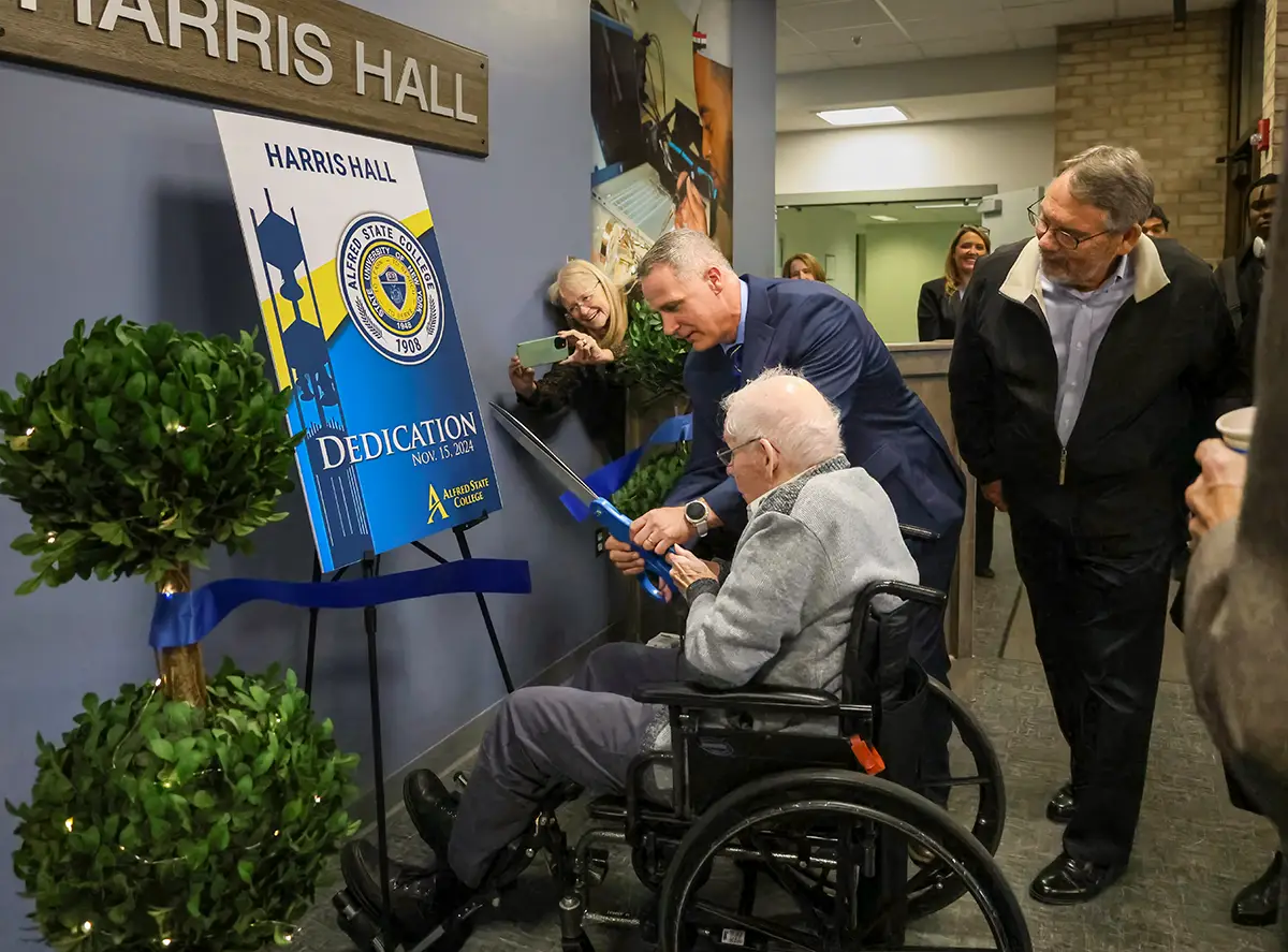 A ribbon-cutting ceremony for Harris Hall, featuring an elderly man in a wheelchair assisted by another man holding large scissors. A sign with ‘Dedication Nov. 15, 2024’ and the Alfred State College logo is displayed. Onlookers, including a woman capturing the moment on her phone, watch the event in a brightly lit hallway.