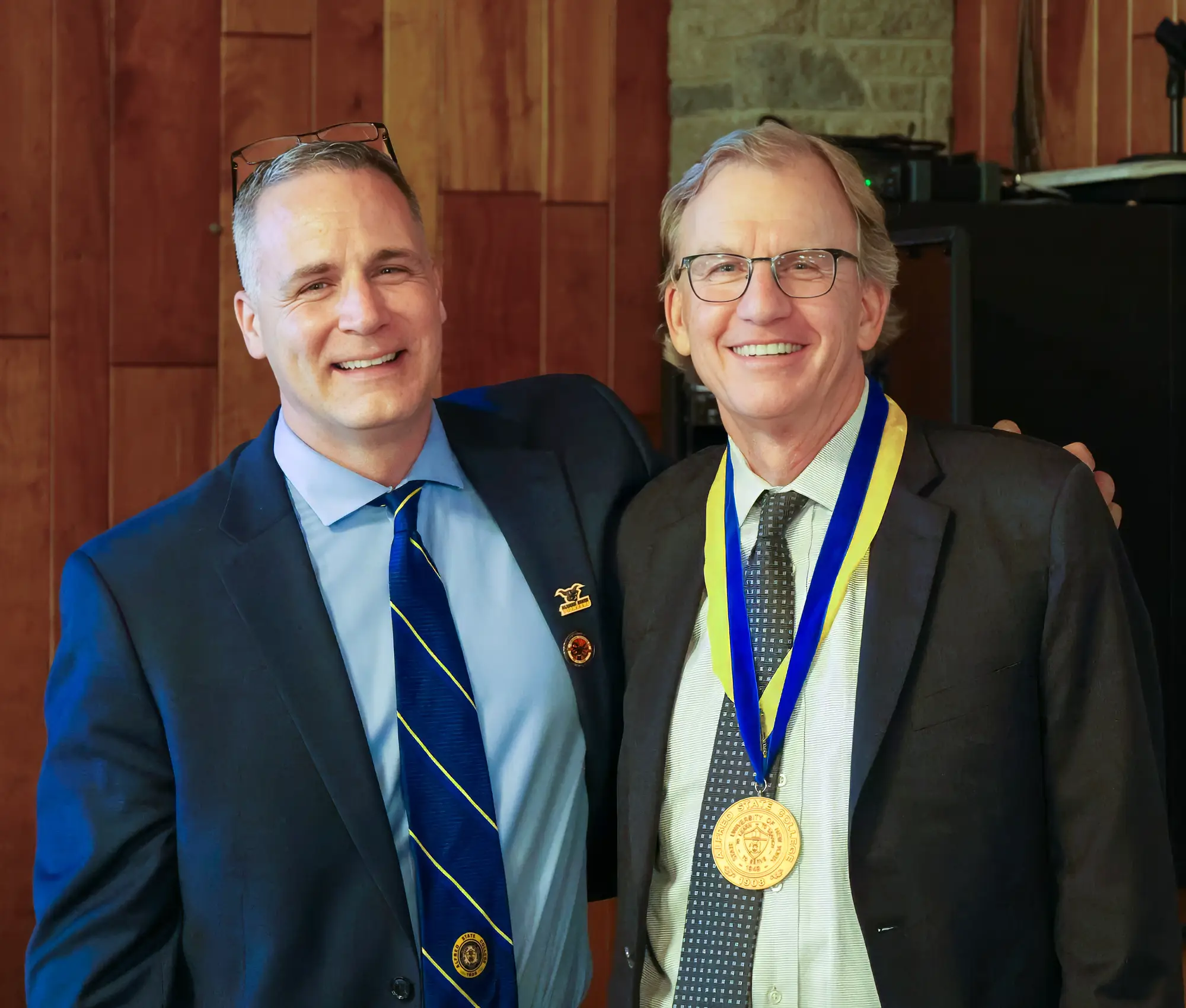 Two men smile for a photo in a formal setting. One wears a suit with a blue striped tie and lapel pins, while the other wears a suit with a medal on a yellow and blue ribbon around his neck. The background features wood paneling and a stone accent.