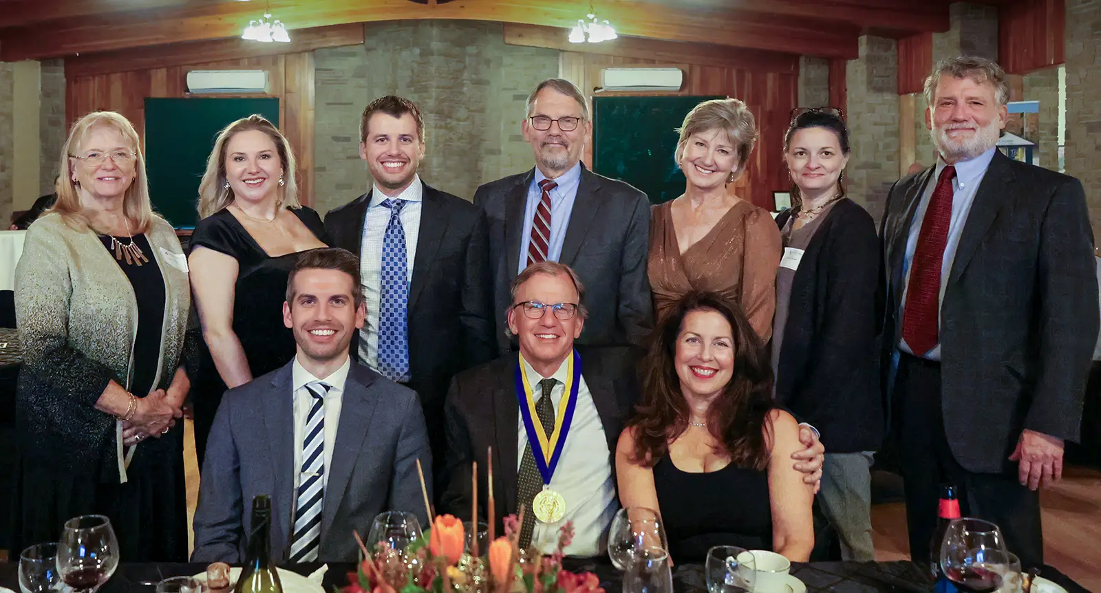 A group of well-dressed individuals poses for a formal photo at a dinner event. The seated individuals are surrounded by others standing behind them, with a beautifully set table featuring wine glasses and floral centerpieces in the foreground. The setting has warm lighting and elegant decor.