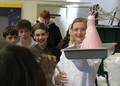 student wearing lab coat showing off a pink volcano-looking object