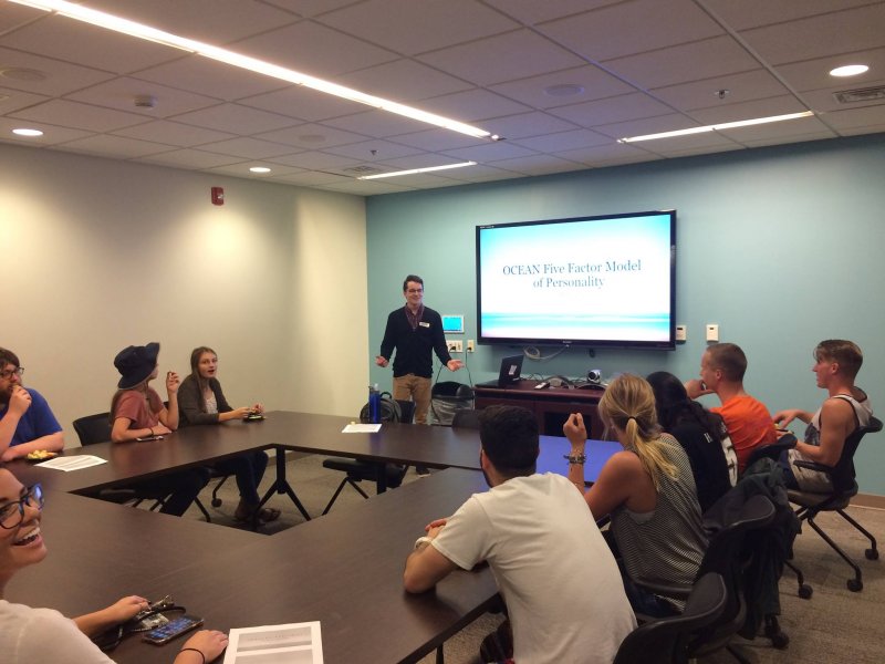 Nine students sit around a square table watching a presentation by another student on "OCEAN Five Factor Model of Personality". The students appear engaged in the presentation.