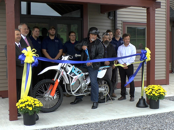 President Sullivan on top of an electric motorcycle  with large scissors, ribbon, several people standing behind him