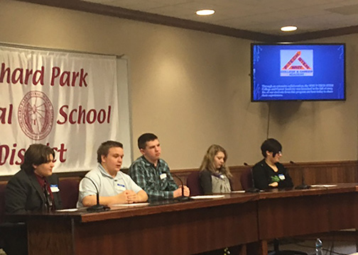 A panel of 5 students sit at a table in front of an audience to answer questions