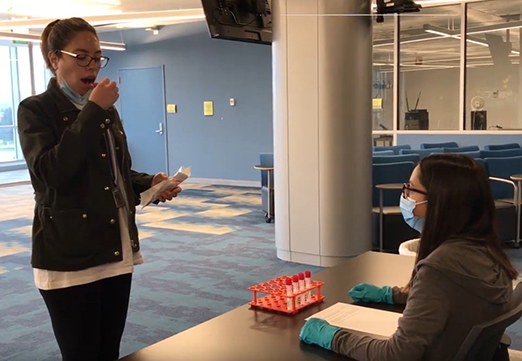 student doing a saliva test in front of another person sitting in a chair behind a table