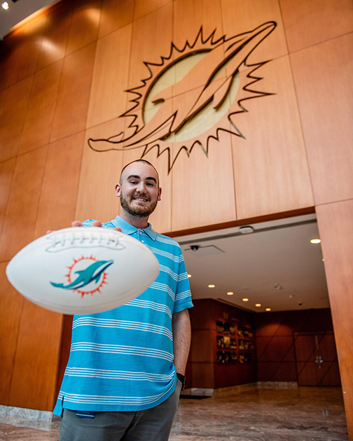 student holding a Miami Dolphins football standing in front of the Dolphin's mascot on the wall