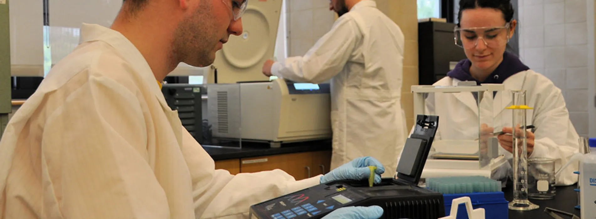 3 students in a lab wearing white coats, blue gloves, using testing equipment