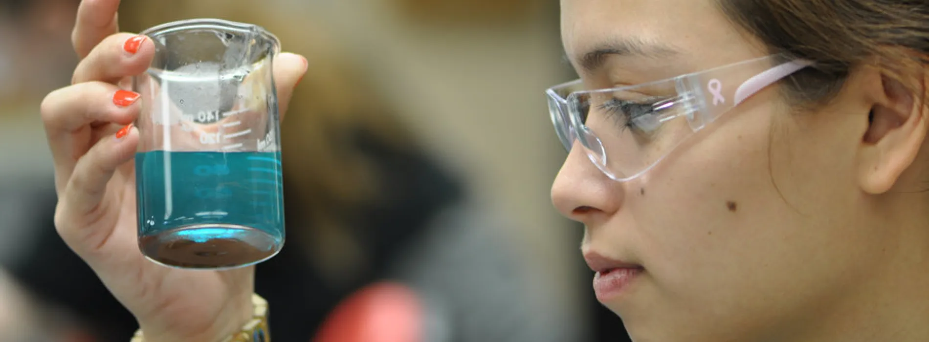 female student wearing safety glasses looking into a beaker with blue liquid 