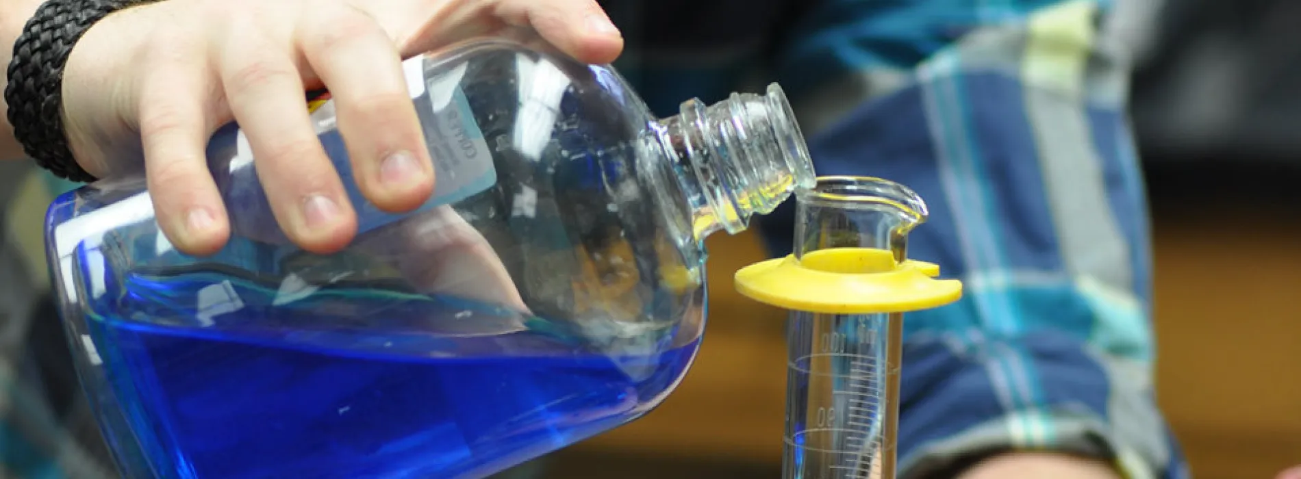 male student hands with cup of blue liquid pouring into a beaker