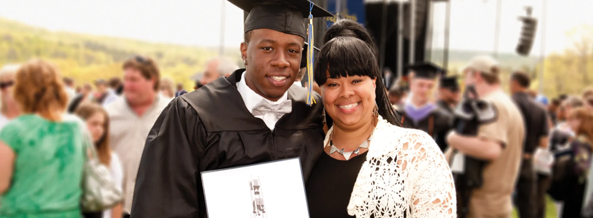 male student wearing commencement cap and gown holding his diploma, with mother