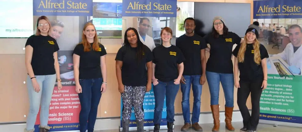 Seven students wearing jeans and blue t-shirts in front of Alfred State posters