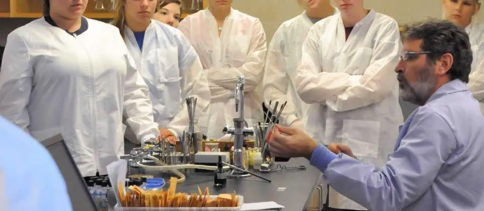 students in white lab coats watching a professor demonstrate something in lab