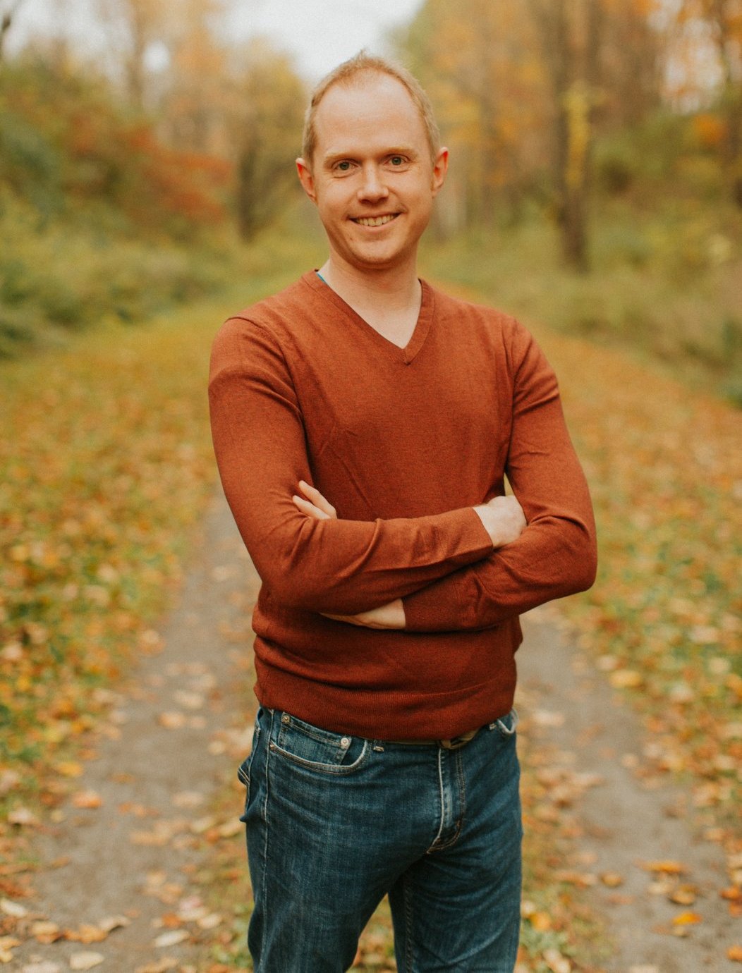 Image includes a picture of Aric Bryant standing in the middle of a road with a fall foliage background.  He has his arms crossed too.  He is also wearing an orange sweater and a pair of blue jeans.