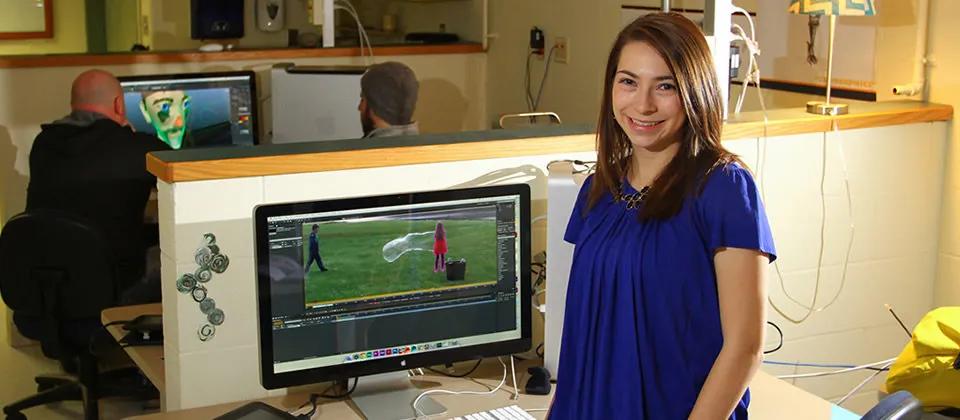 Student posing next to computer with video editing software on screen.