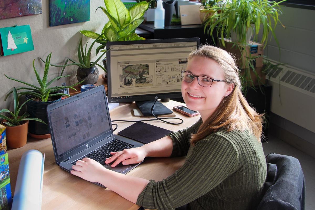 A student smiles while working on a laptop at a desk with design plans displayed on a monitor.