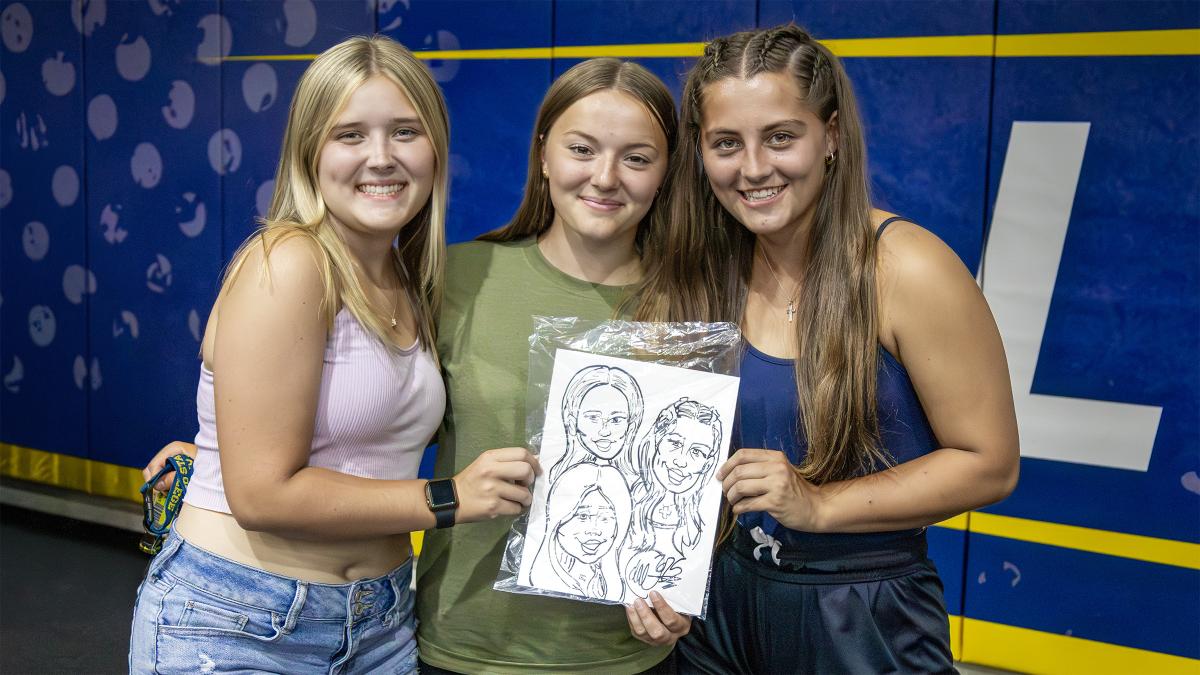 Three young women smile and hold a caricature drawing during a campus event, standing in front of a blue and gold display wall.