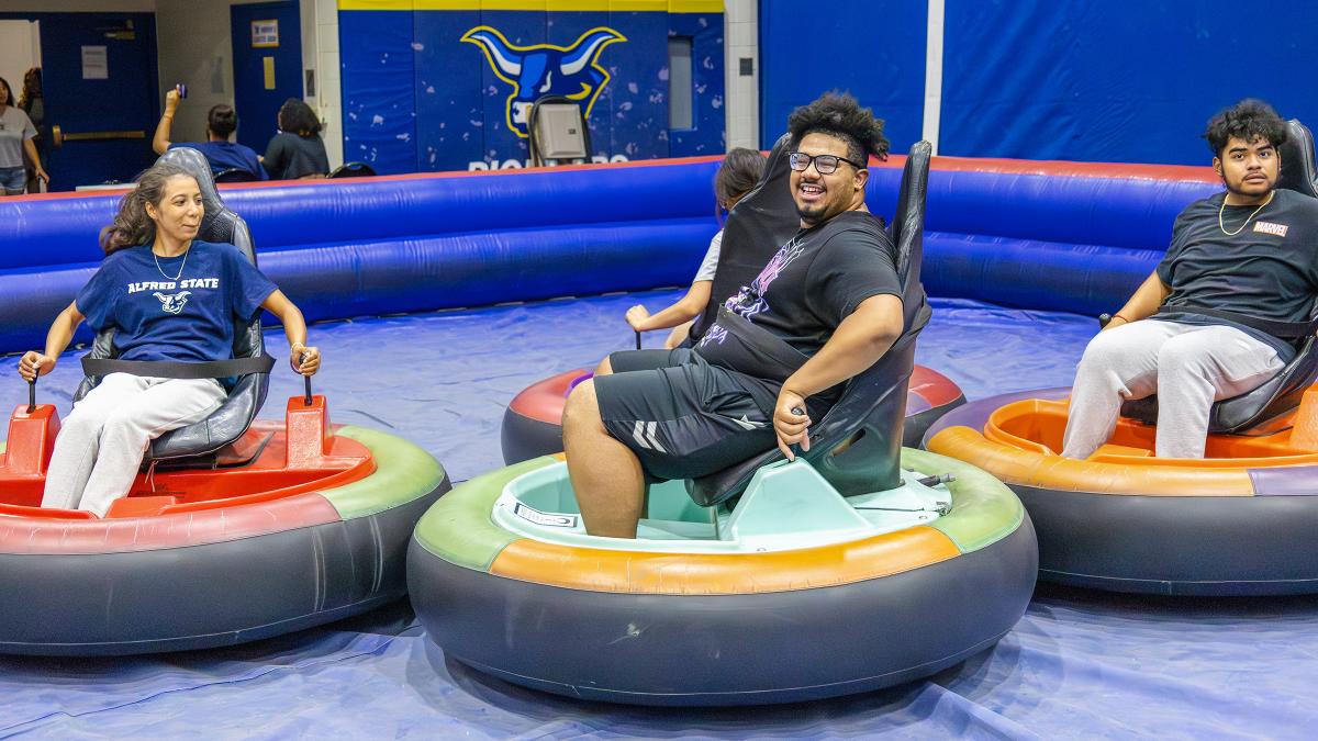 Students ride inflatable bumper cars during a recreational campus event in a gymnasium decorated with Alfred State colors and branding.
