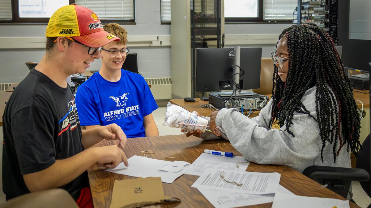 Three students collaborate on a hands on classroom activity, working together at a table with project materials and documents in a computer lab.