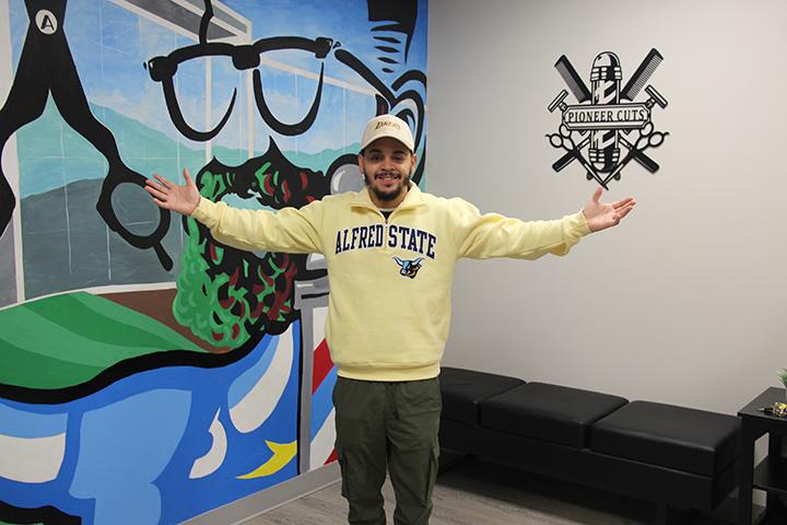 Gabriel Dueño stands in front of the mural in Pioneer Cuts, the new barbershop at Alfred State located in Pioneer Center. 