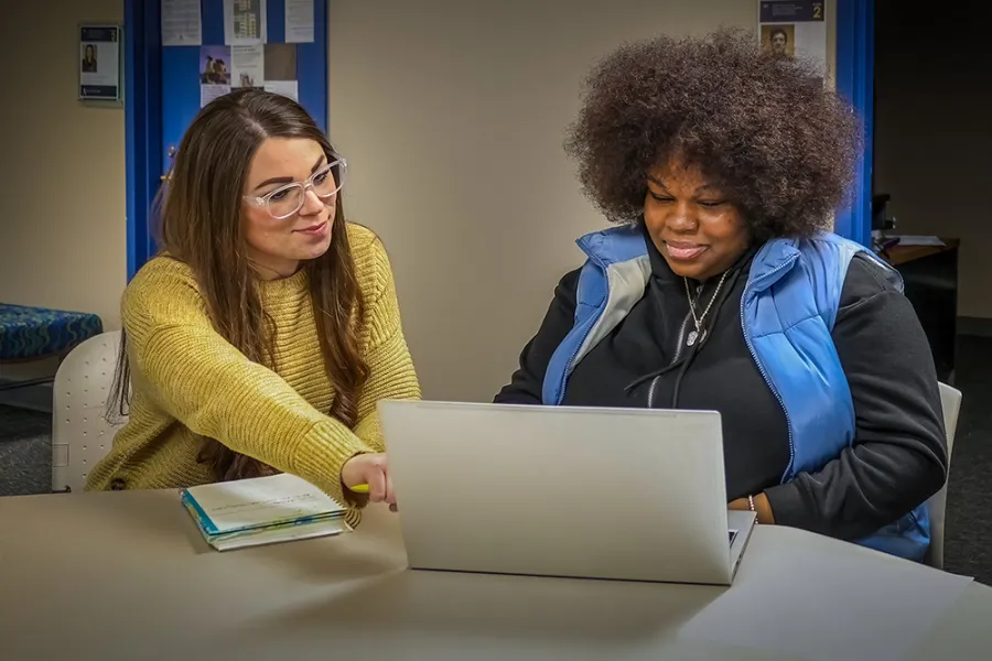 A student working on a laptop while being mentored by a staff member.