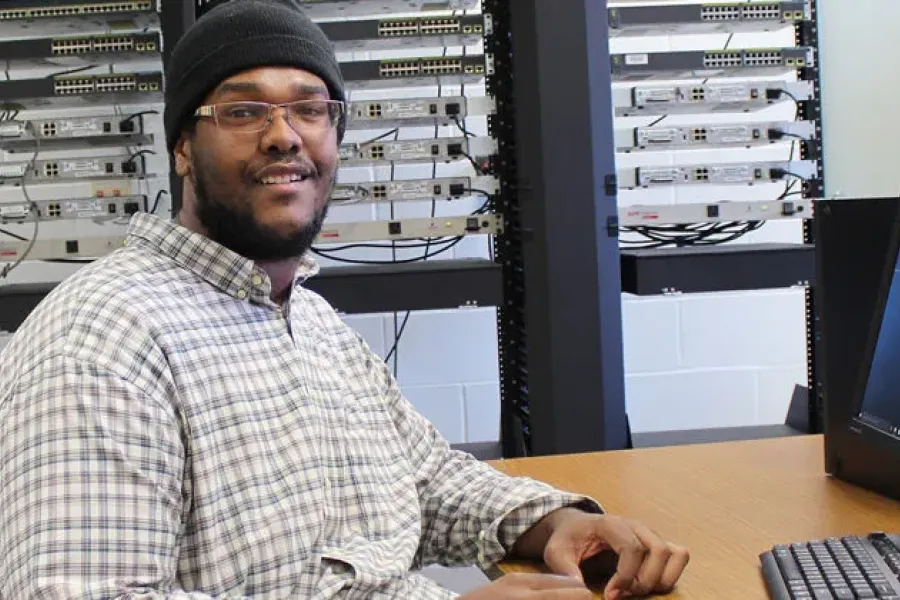 IT student working on computer in classroom with server racks in background.