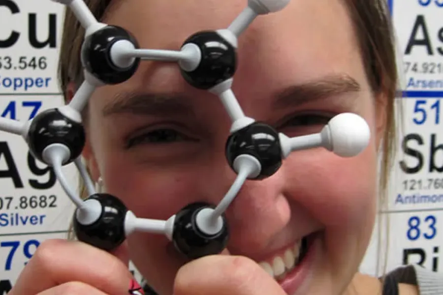 Student holding model of a molecule with periodic table in the background.
