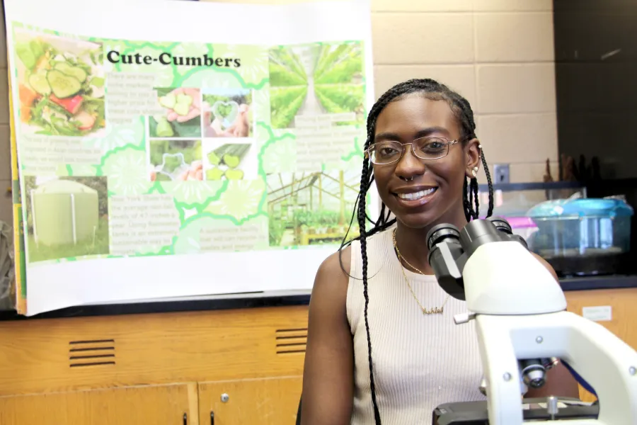 A student sitting in front of a microscope with a research poster on cucumbers in the background