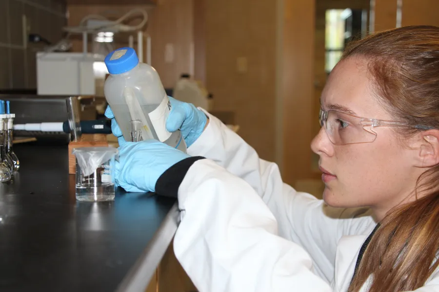 A student measuring a chemical in a beaker while wearing safety gear.