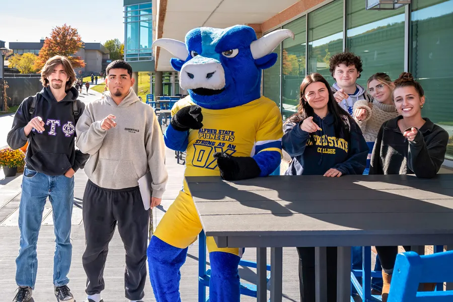 Big Blue, the Alfred State mascot, poses with a group of smiling students outside on campus, all pointing toward the camera.