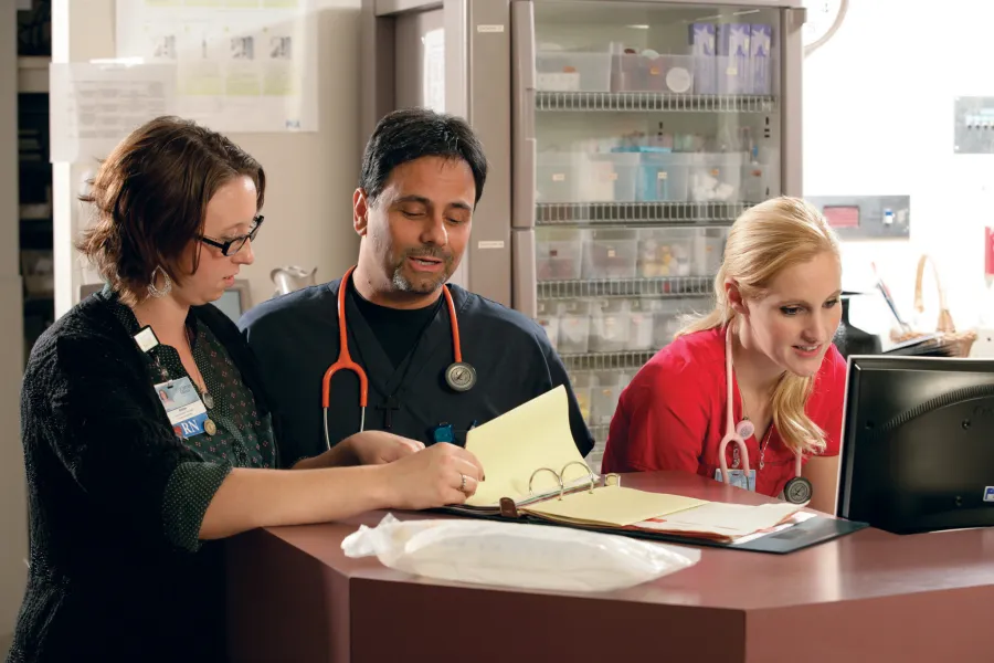 Two medical professionals looking at binder while another is working on a computer in hospital environment.
