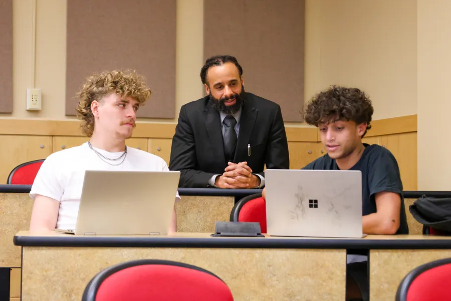 Two students working on laptops in front of a professor who is smiling