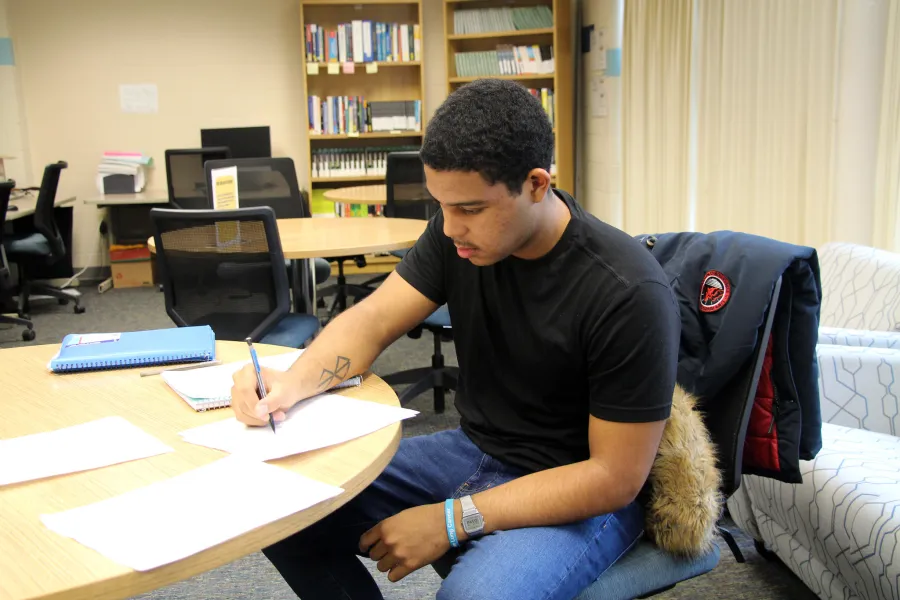 A student sits at a round table in a library or study room, writing on a piece of paper with a pen. He is wearing a black t-shirt, jeans, and a smartwatch, with a blue notebook and additional papers spread out in front of him.