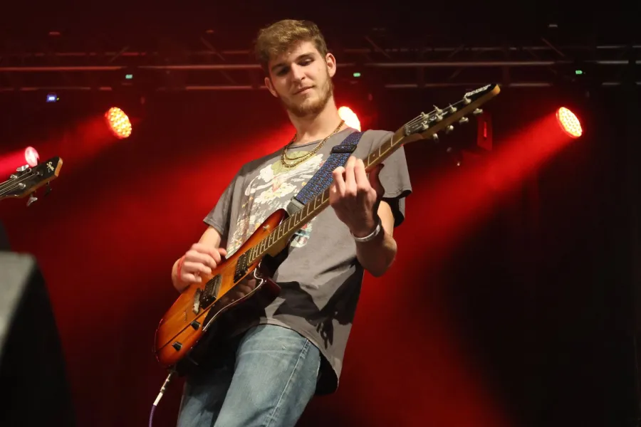 A student plays electric guitar on stage under red concert lights.