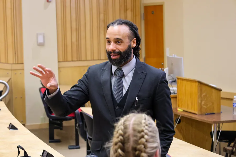 An Alfred State Professor standing at the front of a lecturing hall while wearing a suit and gesturing with with hands to explain a topic.