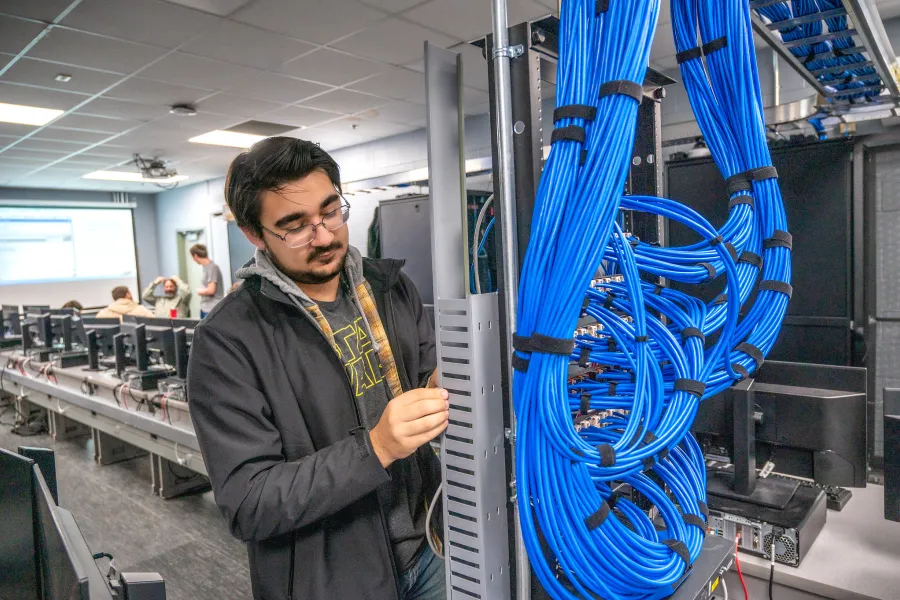 A student maintaining wiring and computer equipment within a cyber security lab.