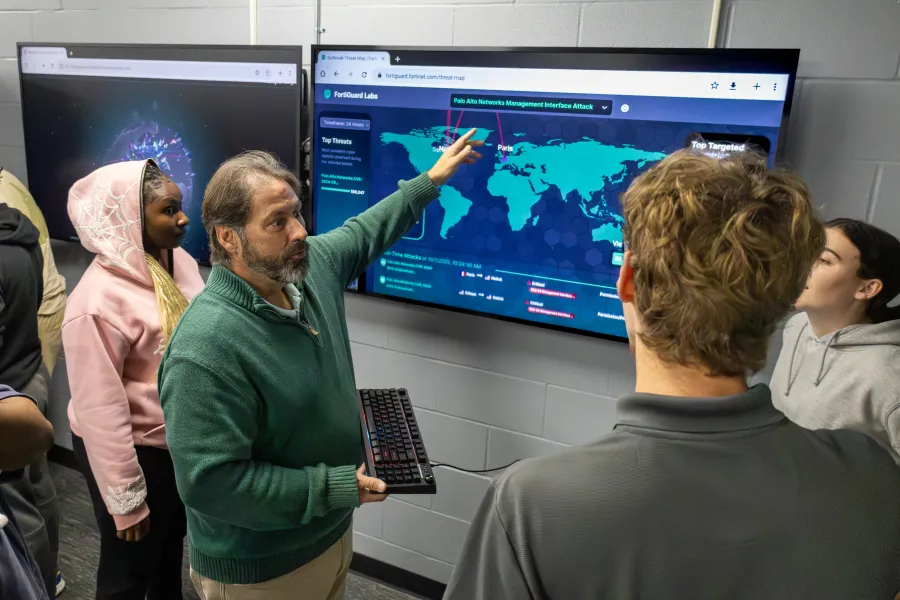 A professor in a cyber security lab pointing at a monitor while students watch.