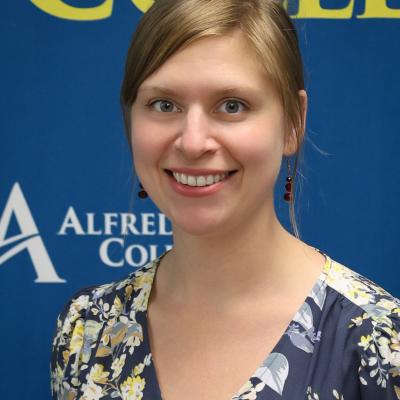 A photo of Amanda Maynes with light brown hair tied back, wearing a textured pink sweater with buttons and coral-colored earrings. The background showcases a serene, blurred natural setting.