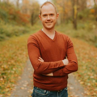 Image includes a picture of Aric Bryant standing in the middle of a road with a fall foliage background.  He has his arms crossed too.  He is also wearing an orange sweater and a pair of blue jeans.